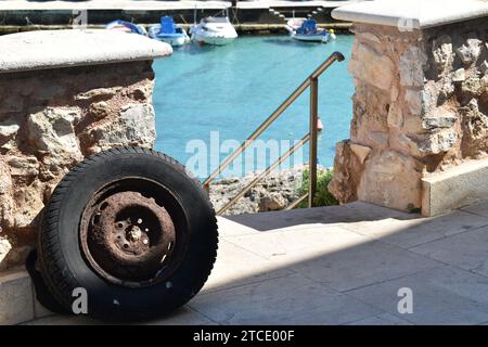Close-up of a rusty tire leaning aginst a rough stone wall with a Mediterranean style marina with boats as blurry background Stock Photo