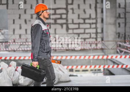 A worker in overalls and a helmet came to a new work site. Builder in uniform carrying toolbox at construction site, copy space Stock Photo