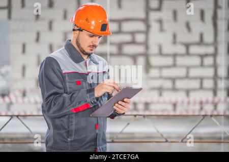 Caucasian construction worker in overalls and helmet using digital tablet on construction site, copy space Stock Photo