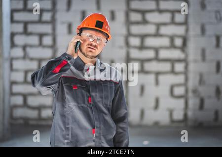 Bearded construction caucasian worker in boilersuit, helmet and protective glusses talking on a smartphone at a construction site Stock Photo
