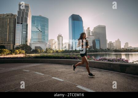 Young Asian woman in sport outfit is running on a track by a lake, with city skyline is in the background. Stock Photo
