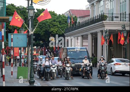 VIETNAM, Hanoi, french quarter, main street with red flag of communist party and Vietnam national flag with yellow star, traffic during rush hour, two-wheeler at crossing Stock Photo