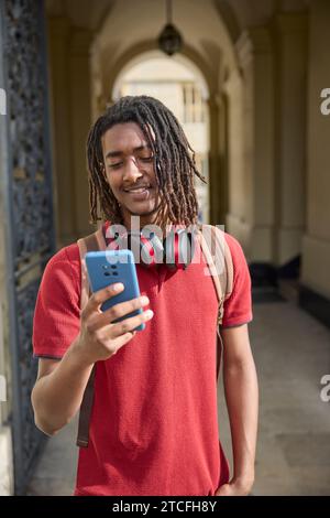 Male Student Wearing Wireless Headphones Checking Messages Or Social Media On Mobile Phone Outside University Building In Oxford UK Stock Photo