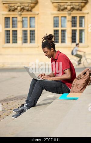 Male Student Working On Laptop Sitting On Steps Outside University Building In Oxford UK Stock Photo