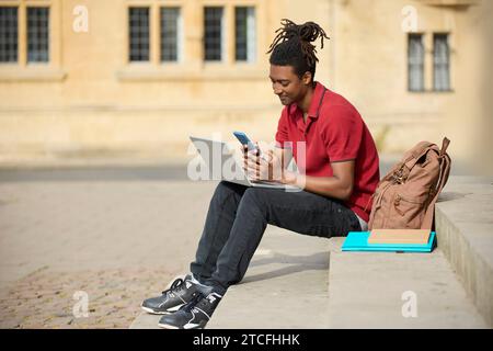 Male Student Working On Laptop Checking Messages Or Social Media On Mobile Phone Sitting On Steps Outside University Building In Oxford UK Stock Photo
