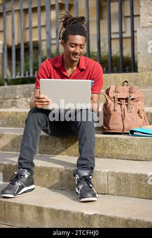 Male Student Working On Laptop Checking Messages Or Social Media On Mobile Phone Sitting On Steps Outside University Building In Oxford UK Stock Photo
