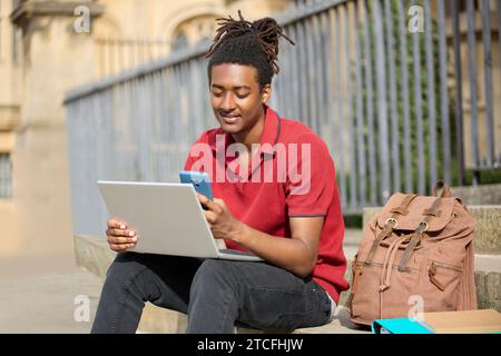 Male Student Working On Laptop Checking Messages Or Social Media On Mobile Phone Sitting On Steps Outside University Building In Oxford UK Stock Photo