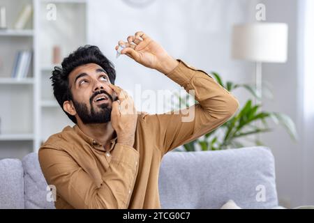 A young Indian man sits at home sick on the sofa and drops medicine in his eyes. Stock Photo