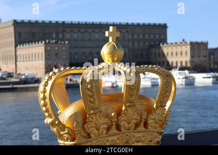 Stockholm Royal Palace. Photo from Skeppsholmsbron with a beautiful golden crown in the foreground. Blue sky. White archipelago boat on calm lake. Stock Photo