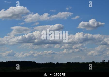 White layered cumulus clouds on a sunny summer day in the blue sky above a dark silhouette of the landscape underneath Stock Photo