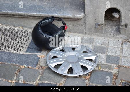 Scratched up grey plastic wheel cover and matte black motorcycle helmet laying on a cobblestone sidewalk in the city centre of Brussels Stock Photo