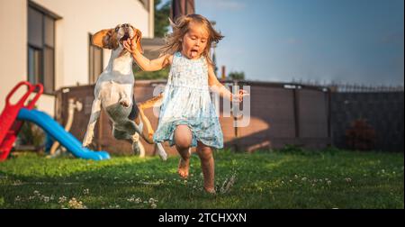 Baby girl running with beagle dog in garden on summer day. Domestic animal with children concept. Dog chasing child with a tennis ball Stock Photo