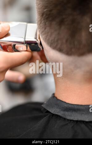 Barber shaves the temple with a cordless trimmer during a short haircut on the sides of the head. Stock Photo
