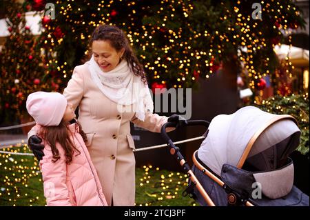 Happy smiling young woman, loving mother looking at her daughter, pushing baby pram, enjoy a Christmas atmosphere outdoors, walking along the beautifu Stock Photo