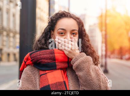 an ethnic curly-haired woman in a jacket and an orange jacket holding her hand to her lips.  Silence is golden concept Stock Photo