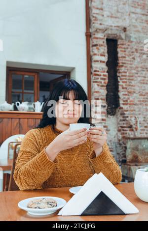 young happy latin woman drinking coffee in cafeteria sitting enjoying alone eating cookie Stock Photo