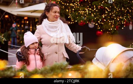 Smiling multi ethnic pretty woman, loving mother hugging her lovely child daughter and pushing baby pram, strolling the Italian street, decorated with Stock Photo