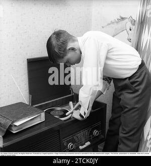 In the 1950s. A teenage boy has put on a gramophone record, a single and carefully places the pickup on the record. It is a radio gramophone, a combined piece of music furniture in which gramophone and radio were combined. A storage album for the discs can be seen on the side. 1958. Kristoffersson ref DB86-5 Stock Photo