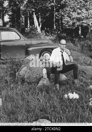 In the 1970s. A man is seen to have stopped after the road, apparently hungry. He has sat on a rock and is eating a sandwich and drinking juice, the packaging is next to it. he is wearing a shirt and tie. The car a Swedish car model Volvo PV 26 July 1971 Stock Photo