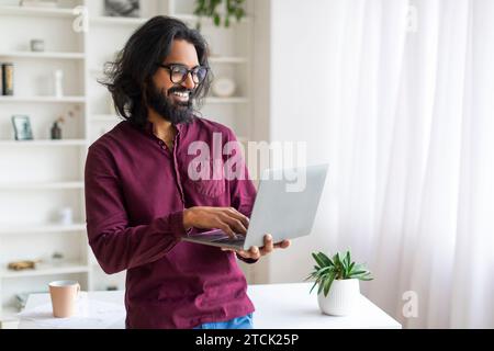 Indian Man Using Laptop While Standing Near Desk At Home, Stock Photo