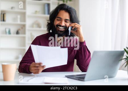 Busy Indian Freelancer Guy Checking Papers And Talking On Cellphone At Home Stock Photo
