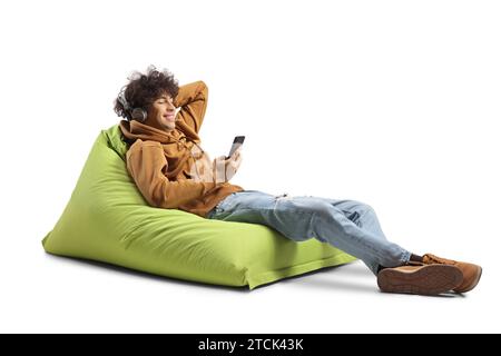 Guy with curly hair sitting on a bean bag armchair and listening to music with headphones and smartphone isolated on white background Stock Photo