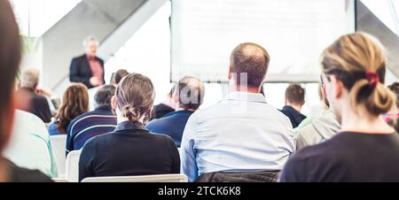 Man giving presentation in lecture hall. Male speeker having talk at public event. Participants listening to lecture. Rear view, focus on people in audience. Stock Photo