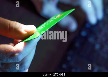 A man, holding a credit card in his hand, dials numbers to pay for online purchases using a computer keyboard Stock Photo