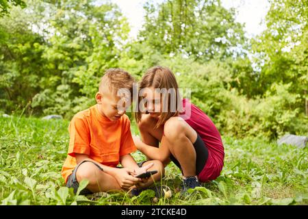 Girl crouching next to boy using smart phone while sitting on grass at park Stock Photo