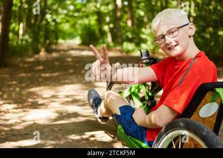Portrait of smiling boy showing peace sign while sitting on recumbent bike at forest Stock Photo