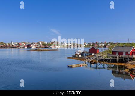 Serene view of the Reine village in Lofoten, Norway, showcasing traditional red rorbu cabins, calm blue waters, and fishing boats under a clear sky Stock Photo