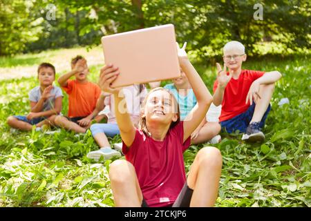 Happy girl gesturing and taking selfie with friends through tablet PC while sitting at park Stock Photo