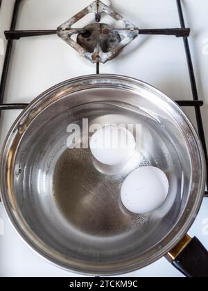 two white chicken eggs are boiling in steel ladle on gas stove close up Stock Photo