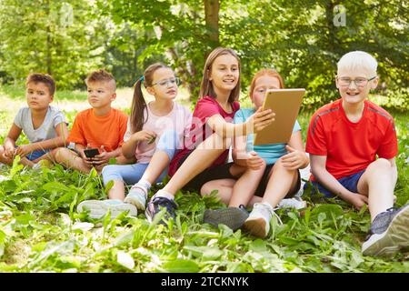 Smiling girl holding tablet PC while sitting with friends on grass at park Stock Photo