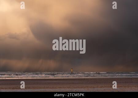 Yellow measuring post in the North Sea, close to the beach, contrasting with dark clouds from which rain falls Stock Photo