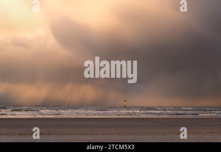 Yellow measuring post in the North Sea, close to the beach, contrasting with dark clouds from which rain falls. Stock Photo