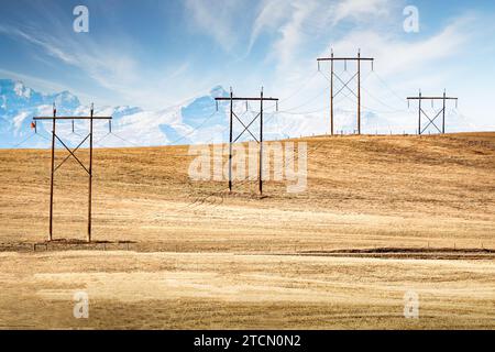 Power lines hanging from large wooden transmission towers over a hilltop overlooking distant mountain tops of the Canadian Rockies in Rocky View Count Stock Photo