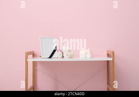 Blank funeral frame, burning candles and flowers on rack near color wall Stock Photo