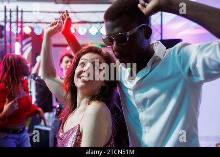 Romantic couple dancing together while relaxing at nightclub party event. Young african american boyfriend and caucasian girlfriend moving on dancefloor, enjoying nightlife Stock Photo