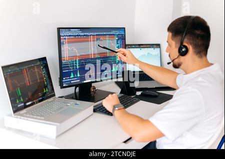 Online trading. Successful male crypto trader sitting at home at his work desk, trading on the stock exchange, holds a pen in hand near monitor and analyzing future of cryptocurrency, assessing risks Stock Photo