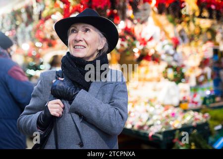 Positive elderly woman in festive mood walking on street Christmas fair Stock Photo