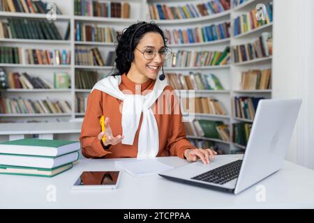 Smiling Indian businesswoman in headset sits at laptop in room, communicates with clients online, works in call center, looks at screen. Stock Photo