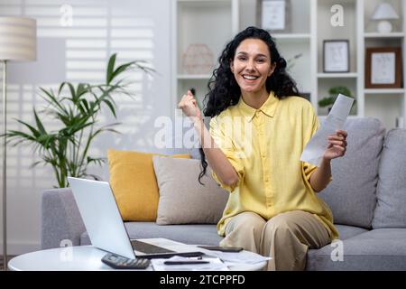 Joyful hispanic businesswoman holding work documents in hands sitting on sofa, happy with good news, success at work, looking at camera. Stock Photo