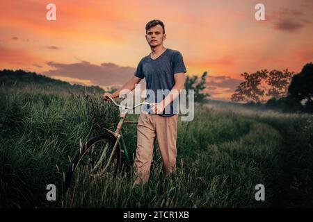 Young man walks with a retro bike along a field road, vintage style in the village at sunset. Sadness and romance in nature Stock Photo