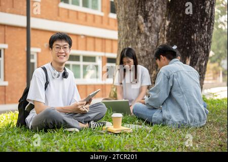 A positive and handsome young Asian male college student in casual outfit with his digital tablet is sitting on grass in a campus park with his friend Stock Photo