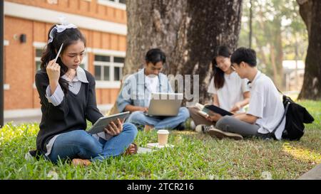 A focused and thoughtful young Asian female college student is using her digital tablet while sitting on grass in a university park with her friends. Stock Photo