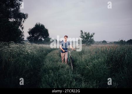 Young man walks with a retro bike along a field road, vintage style in the village at sunset. Sadness and romance in nature Stock Photo