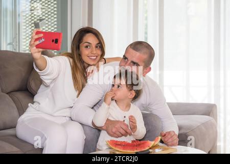 Cute family with baby taking a selfie while eating watermelon in the sofa Stock Photo