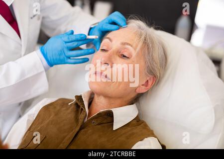 Plastic surgeon hands applying marks on face of elderly female patient Stock Photo