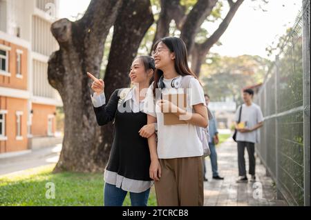 Two cheerful cute Asian female students friends are enjoying chatting while walking to school in the morning together, walking on a footpath, going ho Stock Photo